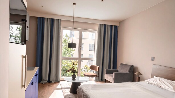 Una habitación de hotel con una cama, una silla, una mesita con una planta y un televisor montado en la pared.