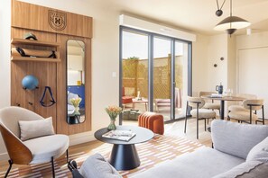 A modern living room with a sofa, armchair, and a coffee table. There is a wooden wardrobe with a mirror and a shelf with a yoga mat and a blue exercise ball. A dining area with wicker chairs and a table is visible through a glass door. - Harvey Chartres — Résidence lifestyle 4★, Restaurant, Bien-Être, Sport & Déconnexion (Chartres)