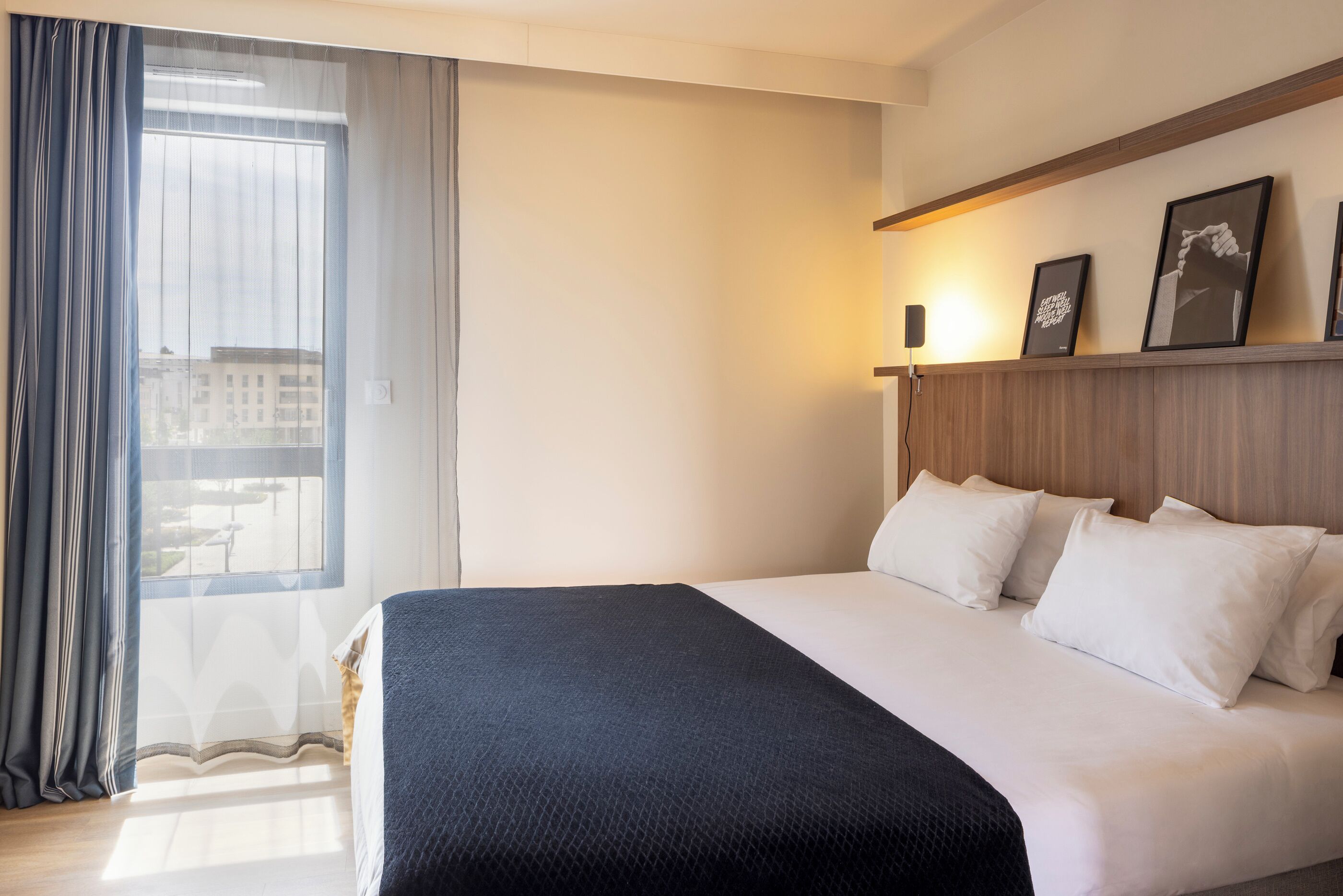 A hotel room with a bed, a window with curtains, a wall-mounted shelf with framed pictures, and a bedside table with a lamp.