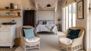 A bedroom with a bed, two wicker chairs, a wardrobe, a shelf with kitchen items, and a window with curtains.