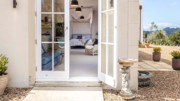 A modern house with a large glass door leading to a bedroom with a bed, a sofa, and a dining area. There are potted plants outside and a stone sculpture in the foreground.