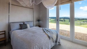 A bedroom with a large bed, a nightstand, a lamp, and a view of the countryside through large windows.