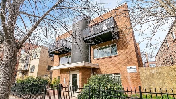 Un bâtiment en briques de deux étages, avec une clôture en métal à l’avant, un panneau indiquant qu’il est interdit d’entrer, et un arbre au premier plan.