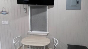 Une pièce avec un téléviseur à écran plat fixé au-dessus d’une fenêtre, une petite table ronde avec deux chaises et un banc sombre.