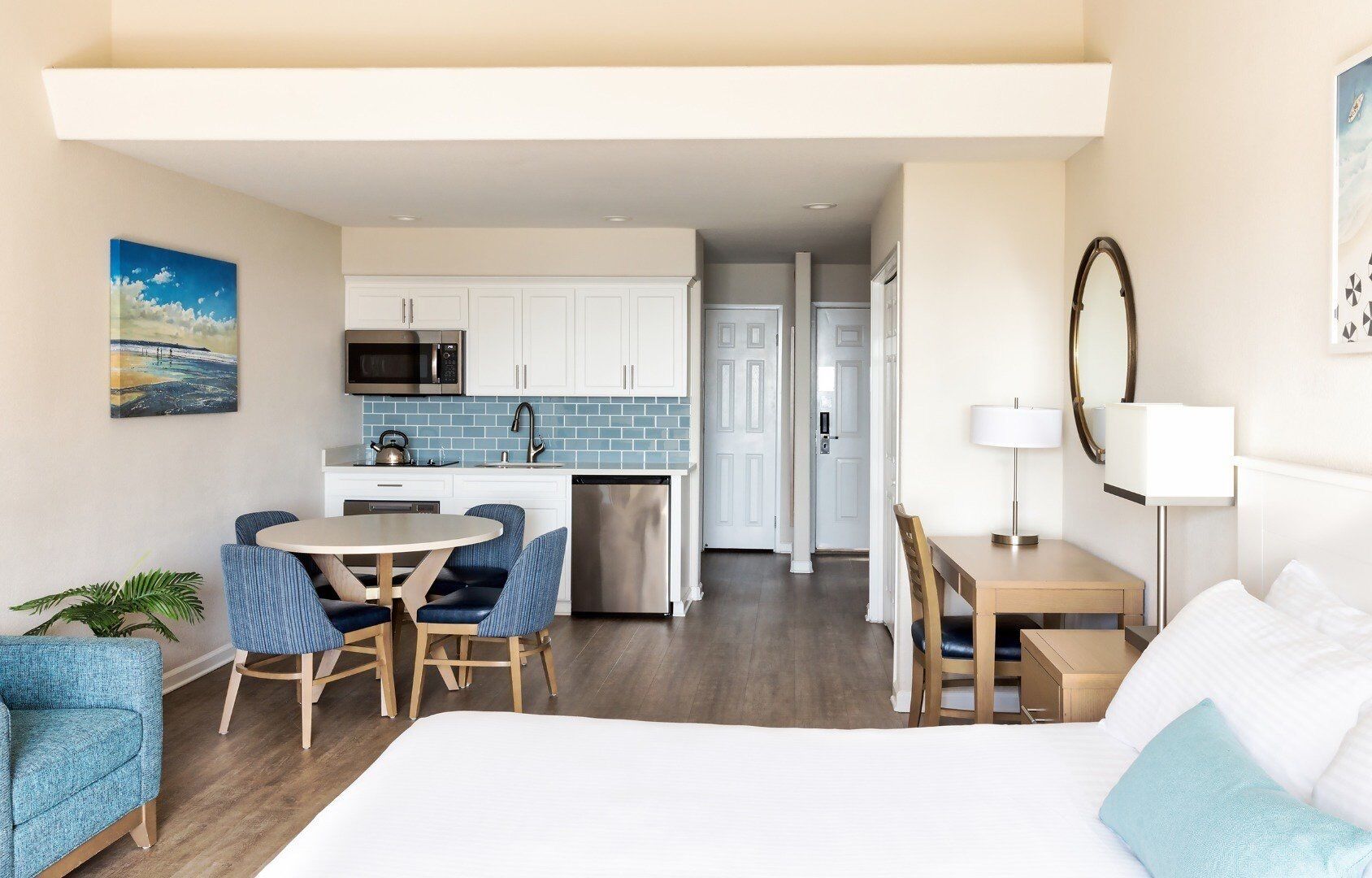 A modern hotel room with a large bed, a dining area with a table and chairs, a kitchen with white cabinets and a blue backsplash, and a view of the hallway.