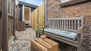 Eine hölzerne Bank mit Kissen, eine Weinflasche und zwei Gläser auf einem Holztisch in einem Innenhof mit Kies.