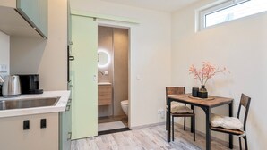 Eine kleine Küche mit Spüle, Wasserkocher und Kaffeemaschine. Ein Esstisch mit Stühlen und eine Vase mit Blumen sind ebenfalls vorhanden. Durch eine offene Tür ist ein WC zu sehen.