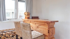 Ett rum med ett skrivbord i trä, flätade stolar, ett fönster med gardiner och en hög med ved.