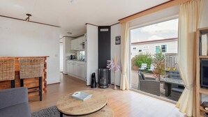 Ett modernt vardagsrum med ett runt soffbord i trä, en grå soffa och en matplats med stolar i rotting. En svart värmare och en krukväxt syns i hörnet.