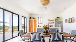 Un'area pranzo moderna con tavolo in legno e sedie a contrasto bianco e nero, una cucina con mobili bianchi e vista sull'esterno attraverso ampie finestre.