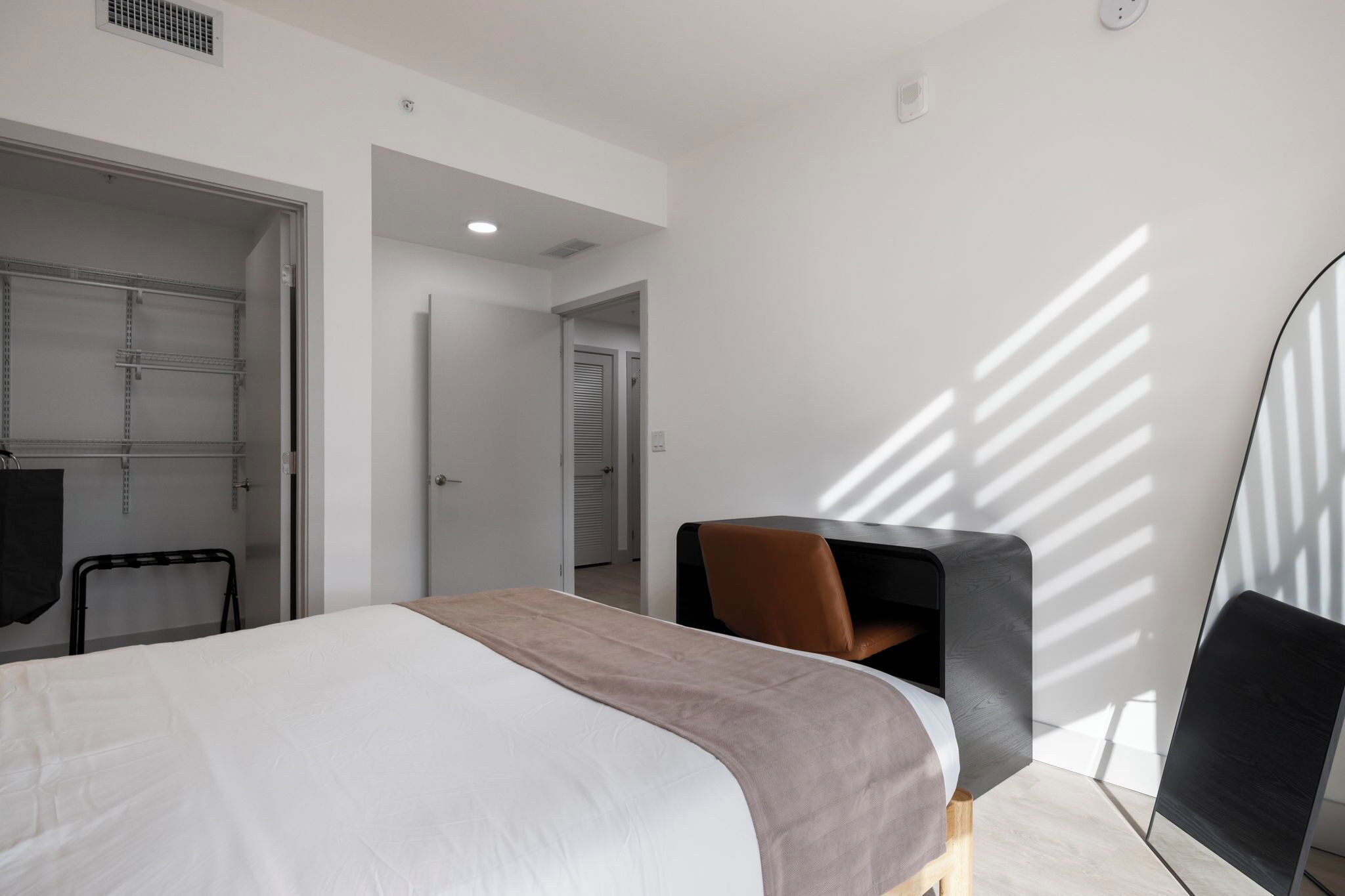 Une chambre d’hôtel moderne, équipée d’un lit, d’un bureau et d’une chaise. On y trouve également une armoire et une porte donnant sur une autre pièce.