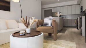 A modern living room with a sofa, a round coffee table with a vase, a floor lamp, and a kitchen in the background.