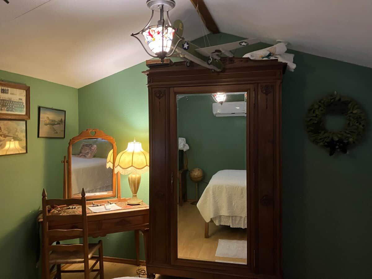 Une pièce comprenant un bureau en bois, une chaise, une lampe, un lit recouvert d’une courtepointe blanche, un miroir et un ventilateur de plafond.