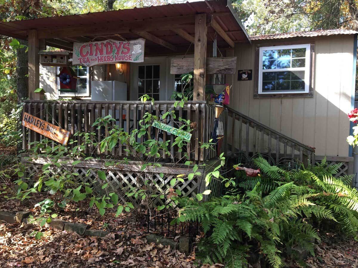 A rustic building with a wooden porch, signs for 'CINDY'S TEA AND HERBS HOUSE' and 'HARLEY'S BAR', and a welcoming 'WELCOME' sign.