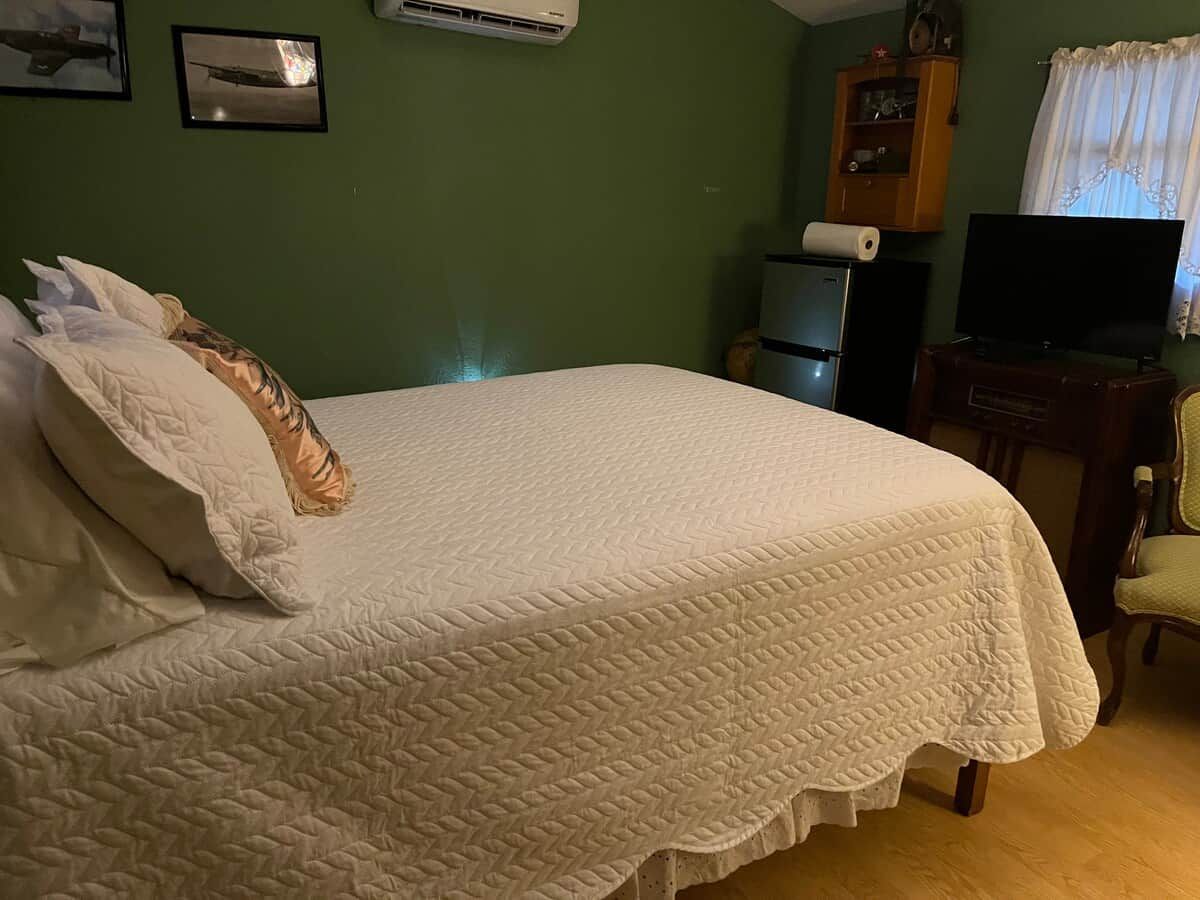 Una cama bien hecha con colcha blanca, un televisor, un pequeño mueble y una silla en una habitación con paredes verdes.