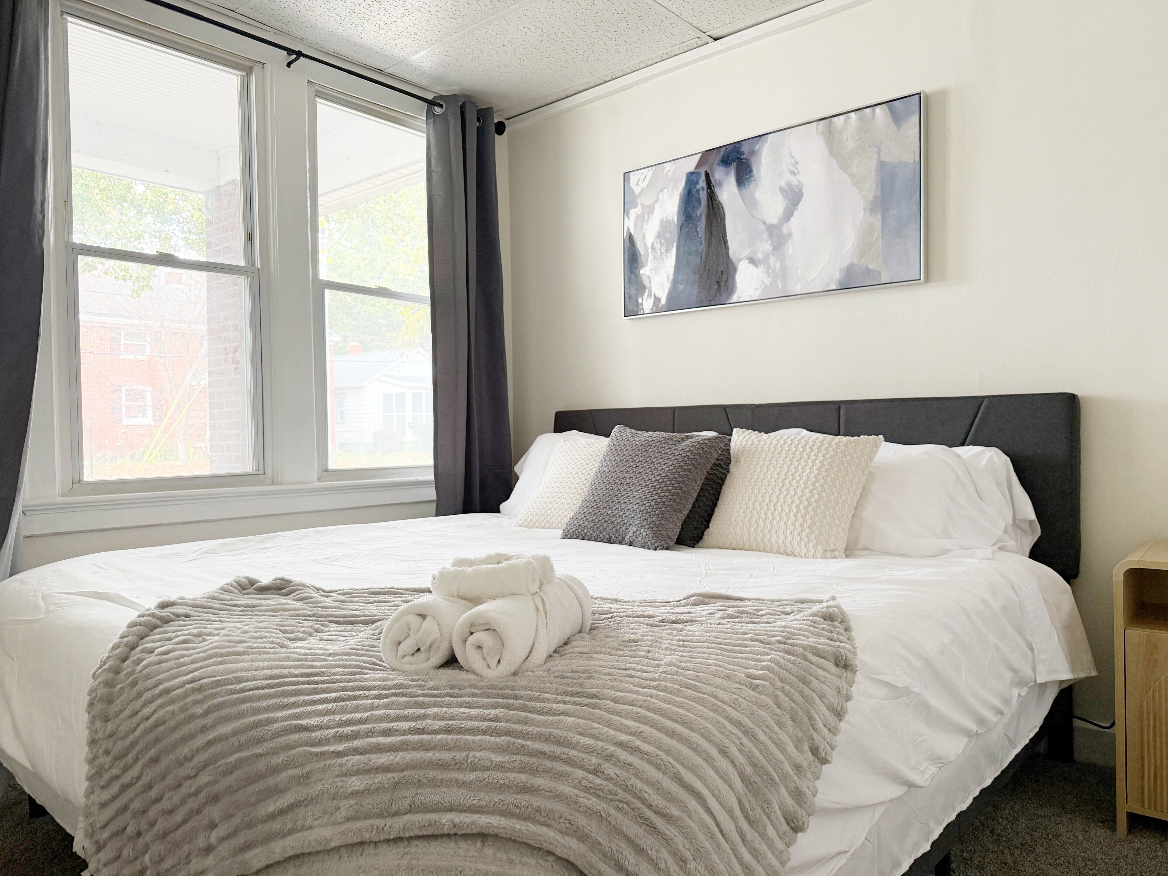 A neatly made bed with a grey blanket and rolled white towels, a grey headboard, and a painting on the wall.