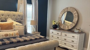 A bedroom with a tufted headboard, a bedside table with a lamp, a dresser with drawers, a round mirror, and a patterned rug. - Cozy 1-bedroom studio in gorgeous El Paso (El Paso)