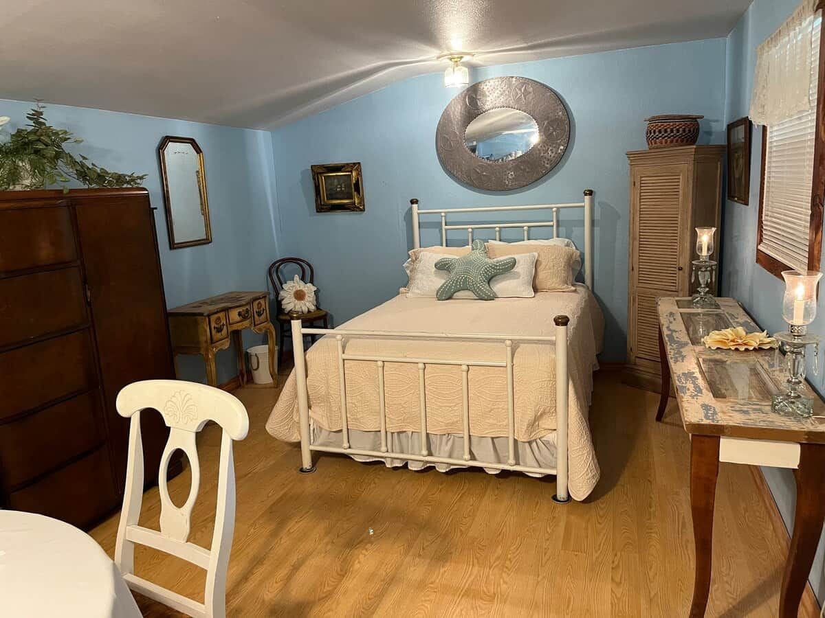 A bedroom with a bed, a dresser, a chair, a table with a lamp, and a mirror on the wall.