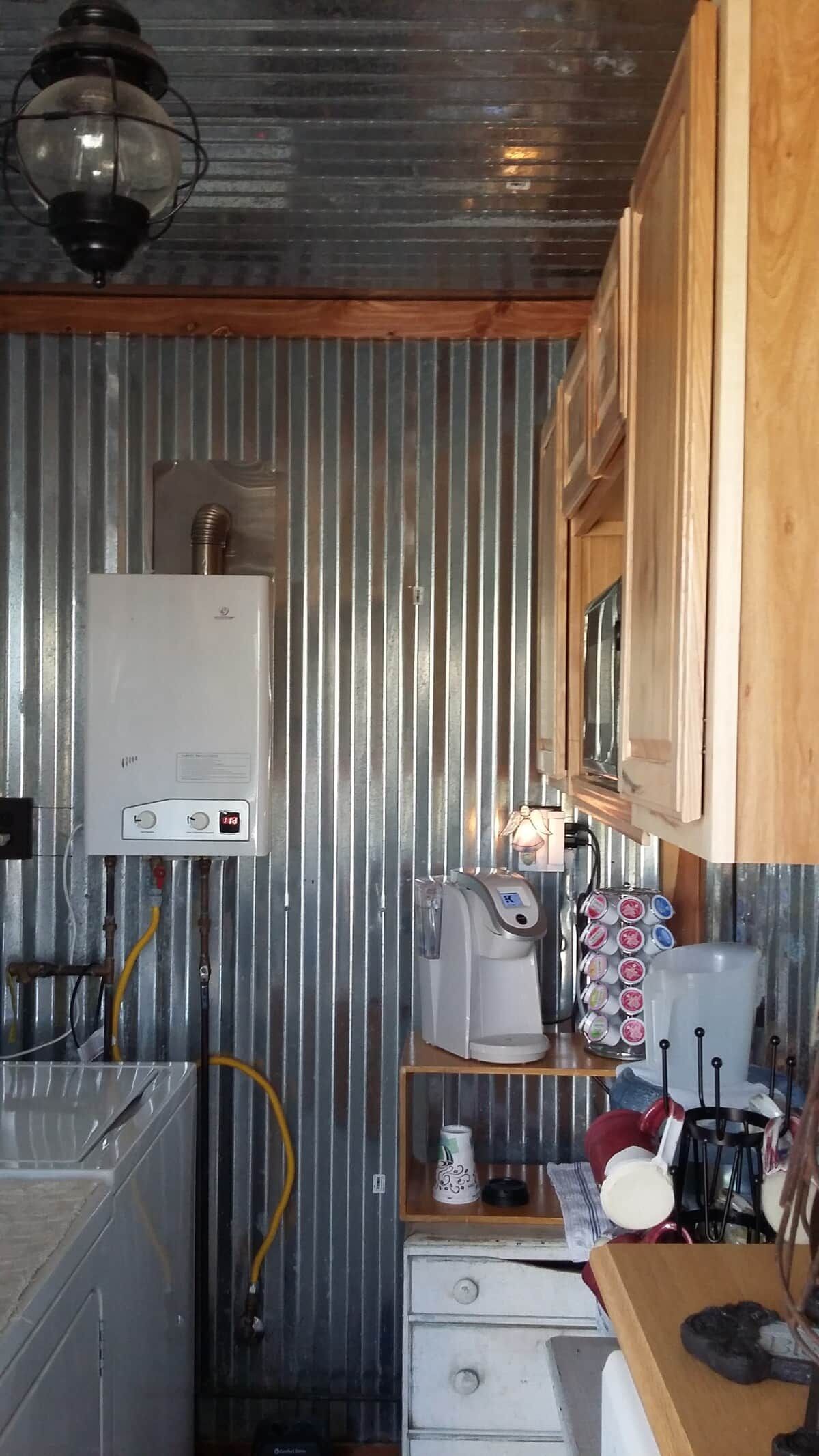 A compact kitchen with a water heater, coffee maker, and a shelf with various items.