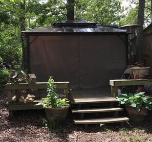 A covered outdoor seating area with a wooden deck and steps leading up to it, surrounded by trees. - The BoatHouse (Tyler)