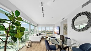 A modern living room with a glass dining table, blue chairs, a white sofa, and a large window offering a city view. - Anse Lasio' Penthouse Panoramic Ocean View Rooftop BBQ, Pool and Gym (Santa Monica)