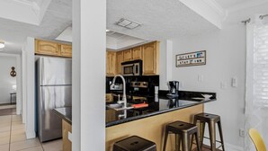 A modern kitchen with stainless steel appliances, wooden cabinets, and a bar stool area. - Turtle Tide at Del Mar - Charming Condo w/ Heated Pool, Beach Equipment, & Steps To The Beach (Indian Rocks Beach)