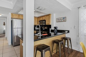 A modern kitchen with stainless steel appliances, wooden cabinets, and a bar stool area. - Turtle Tide at Del Mar - Charming Condo w/ Heated Pool, Beach Equipment, & Steps To The Beach (Indian Rocks Beach)