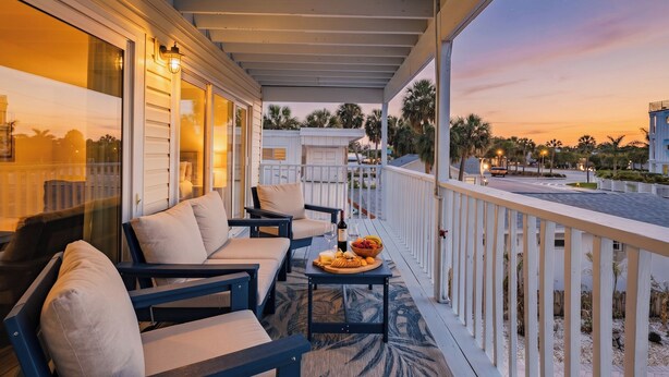 A covered outdoor seating area with a table set for dining, a railing, and a view of the street and palm trees. - Turtle Tide at Del Mar - Charming Condo w/ Heated Pool, Beach Equipment, & Steps To The Beach (Indian Rocks Beach)