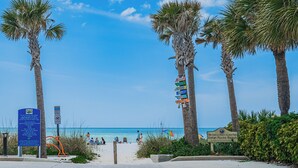 A beach entrance with palm trees, a sign for 19th Avenue Beach Access Park, and a beach with people. - Turtle Tide at Del Mar - Charming Condo w/ Heated Pool, Beach Equipment, & Steps To The Beach (Indian Rocks Beach)