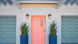 A house with a pink door, two potted plants, and two garage doors. - Turtle Tide at Del Mar - Charming Condo w/ Heated Pool, Beach Equipment, & Steps To The Beach (Indian Rocks Beach)