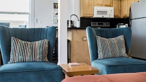 Uma sala de estar moderna com dois poltronas azuis e uma pequena mesa de madeira, área de cozinha com armários e eletrodomésticos de madeira, e vista para o exterior através de uma janela.