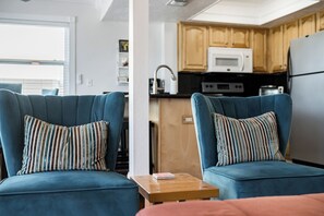 A modern living room with two blue armchairs and a small wooden table, a kitchen area with wooden cabinets and appliances, and a view of the outdoors through a window. - Dolphin Drift at Del Mar - Charming Condo w/ Heated Pool, Beach Equipment, & Steps To The Beach (Indian Rocks Beach)