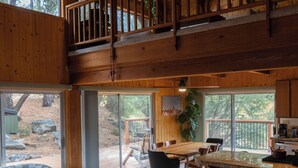 Uma cabana de madeira espaçosa com uma grande sala de jantar, cozinha e varanda com vista para uma área arborizada.