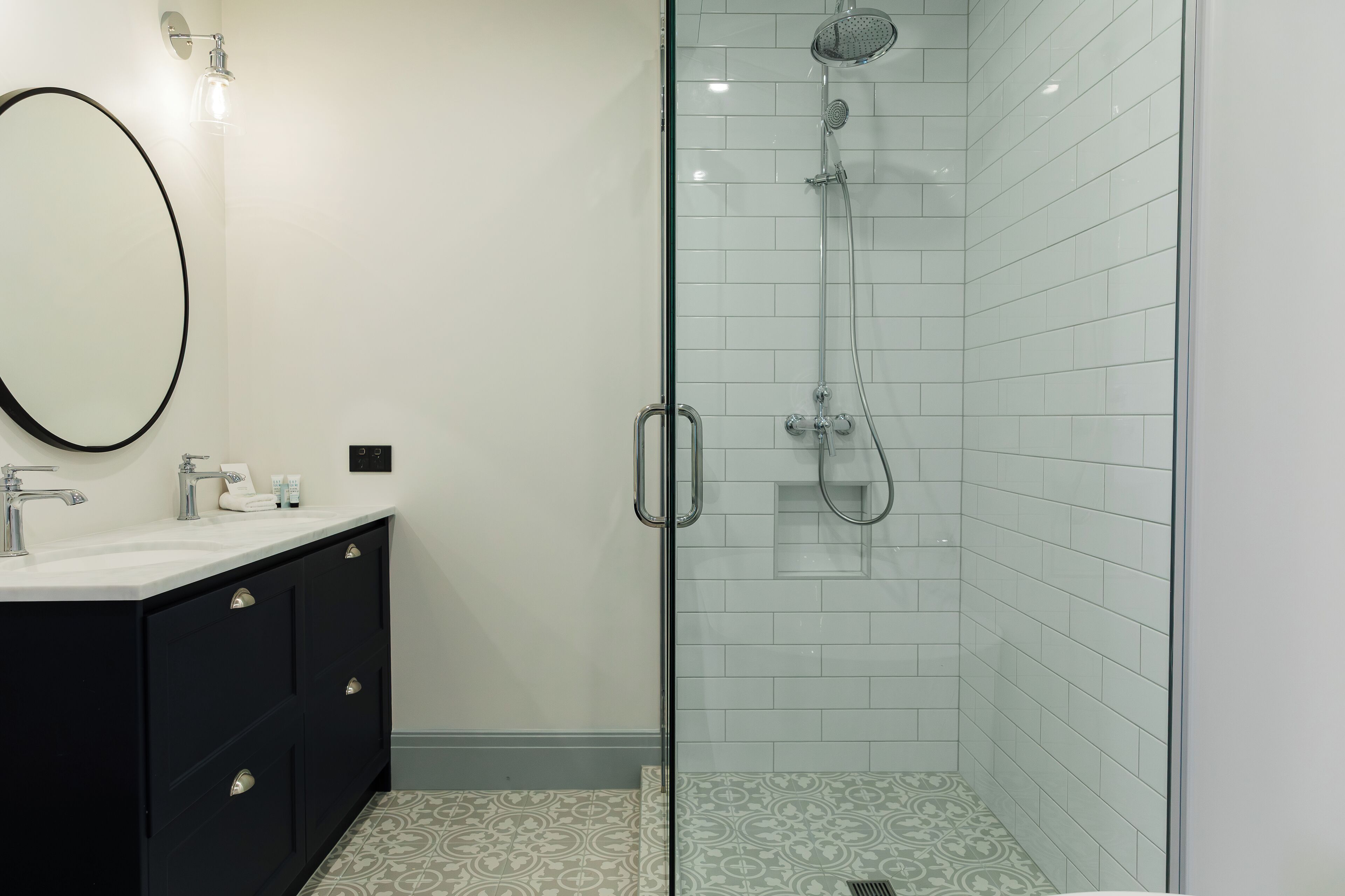 Un baño moderno con cabina de ducha de vidrio, tocador negro con espejo y baldosas de suelo estampadas.