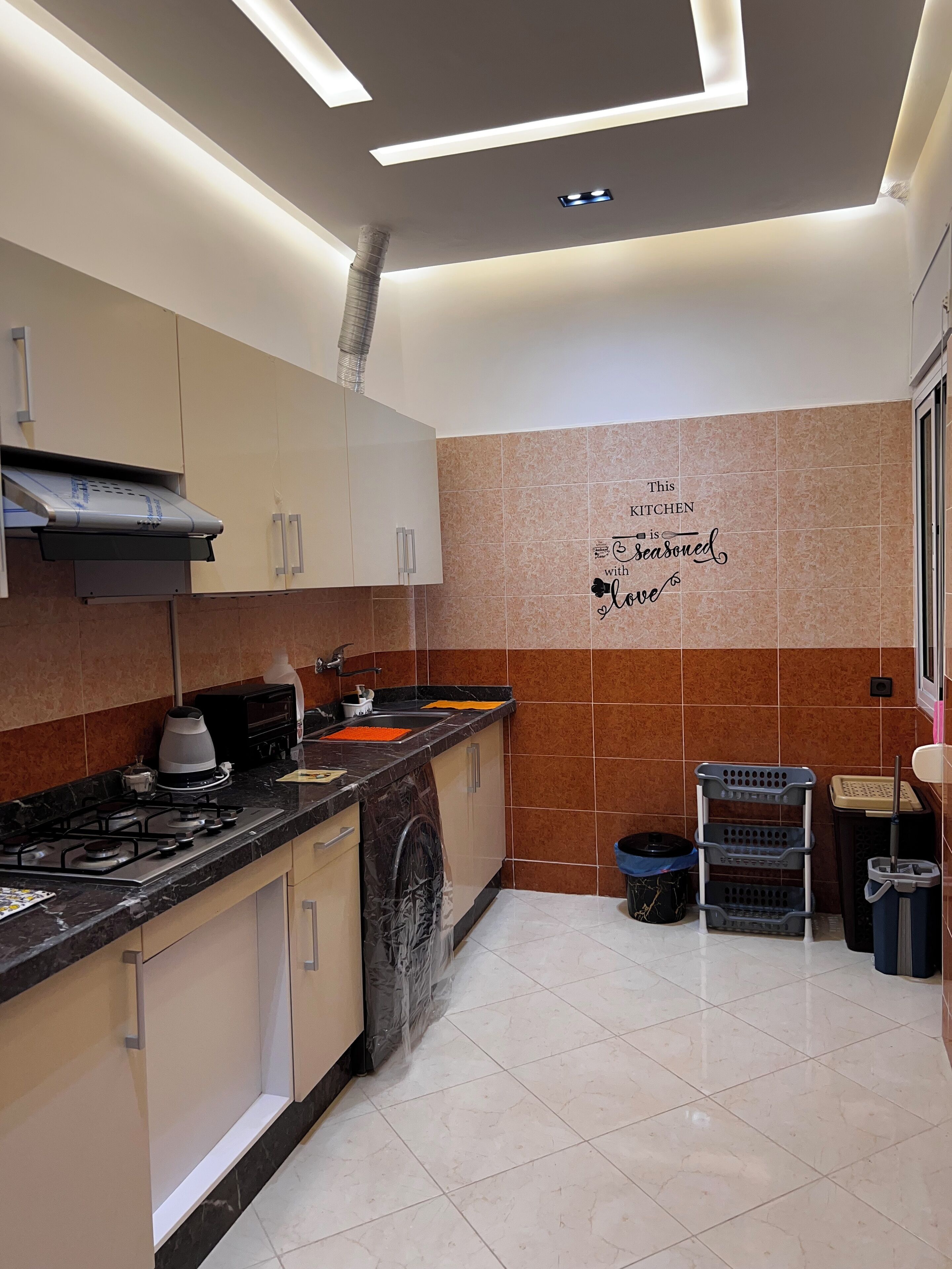A modern kitchen with a gas stove, oven, and a built-in range hood. There are cabinets, a countertop, and a wall-mounted microwave. The floor is tiled, and there is a trash bin and a laundry basket in the corner.