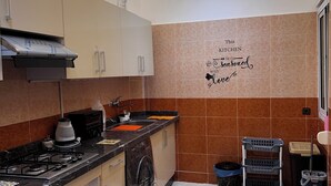A modern kitchen with a gas stove, oven, and a built-in range hood. There are cabinets, a countertop, and a wall-mounted microwave. The floor is tiled, and there is a trash bin and a laundry basket in the corner. - Belmar Martil - Chic and close to the sea (Martil)
