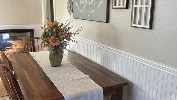 A dining room with a long wooden table, white tablecloth, and a vase of flowers. There are two arched windows on the wall and a framed quote above the windows.