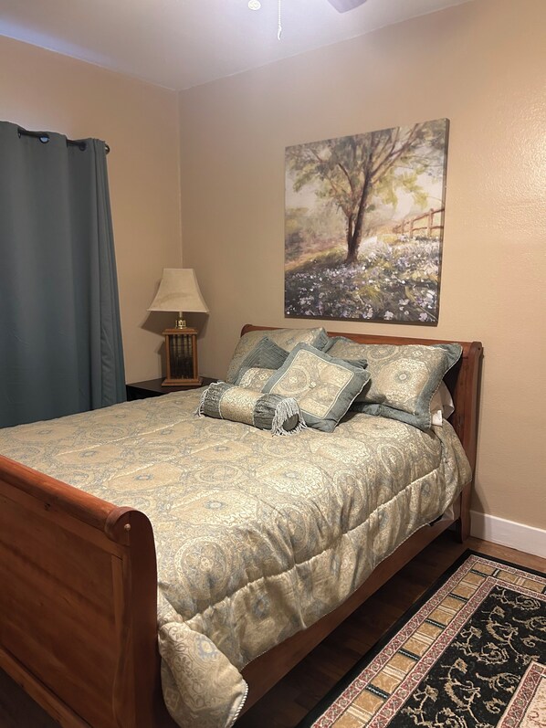 A neatly made bed with a patterned bedspread and decorative pillows, a painting on the wall, and a wooden nightstand with a lamp.