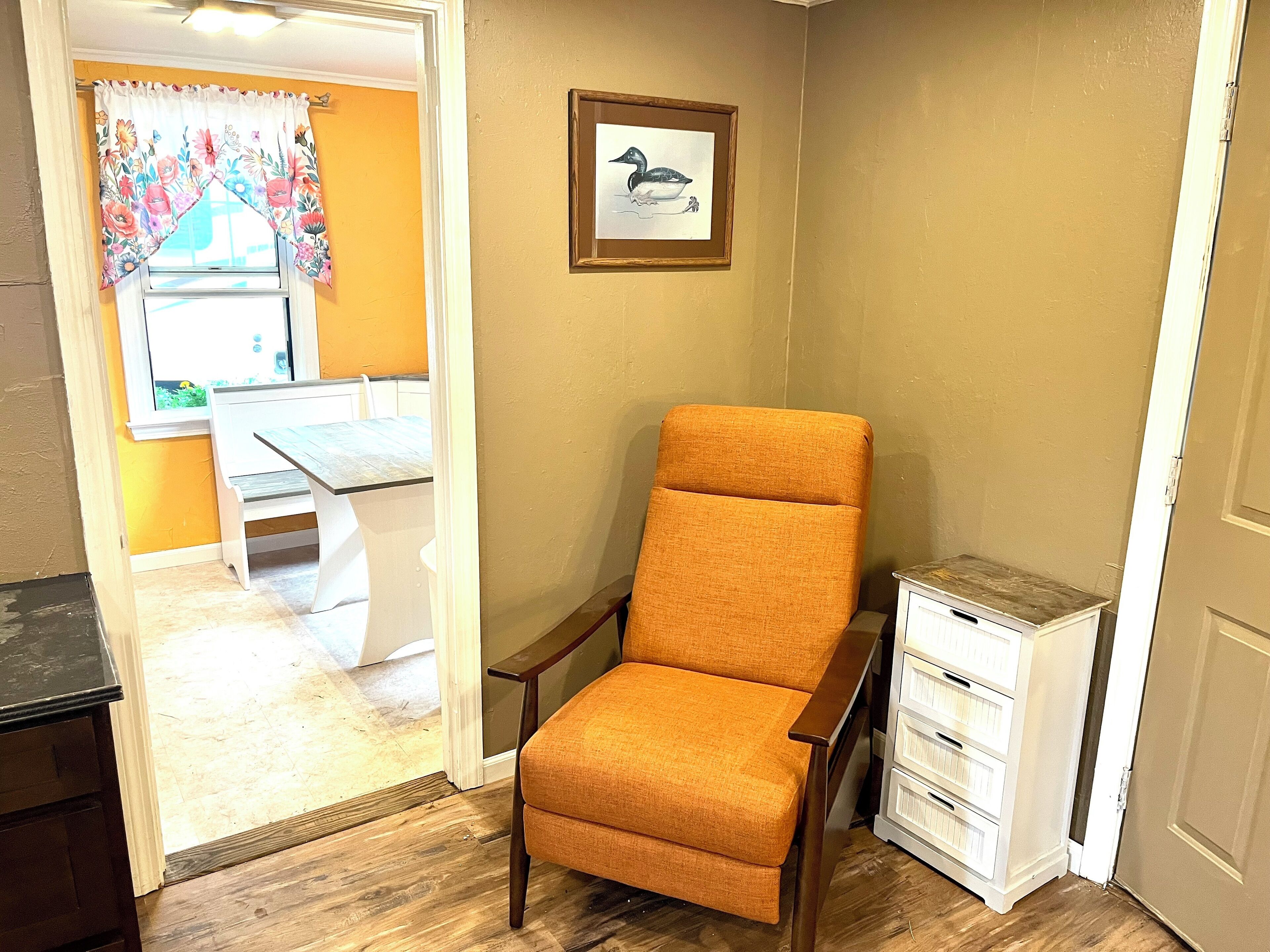 A room with a wooden floor, a white table with chairs, a framed picture on the wall, and a small white cabinet.