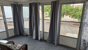 Ein Zimmer mit einem großen Fenster, grauen Vorhängen, einem Korbstuhl mit floralem Kissen und einem grauen Teppich.