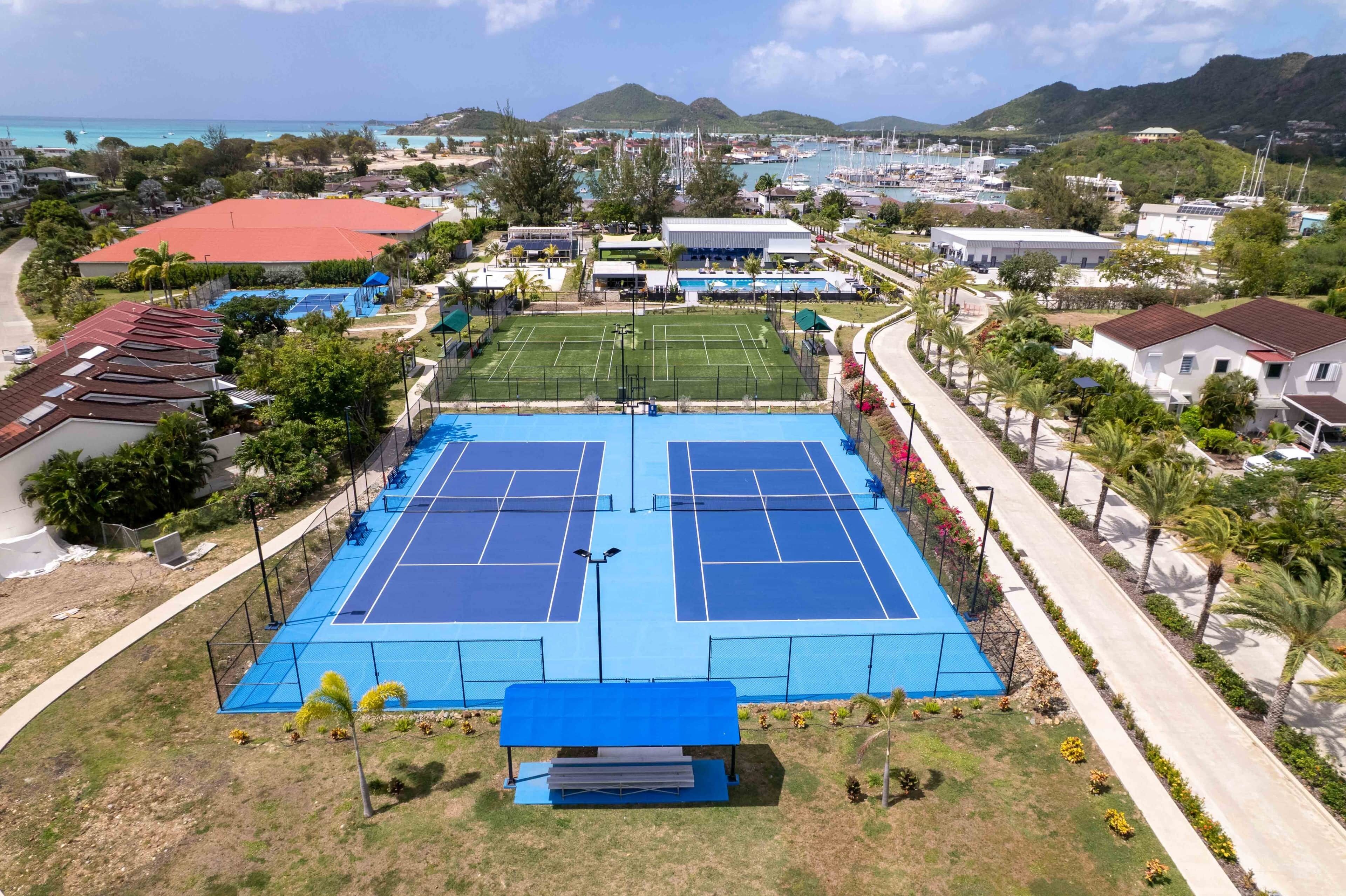 A tennis court with a net, surrounded by a fence, and a building with a red roof in the background.