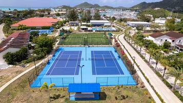 A tennis court with a net, surrounded by a fence, and a building with a red roof in the background.