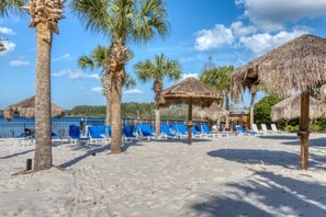 Ein Strand mit Palmen, strohgedeckten Hütten und blauen Liegestühlen.