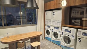 Een wasserette met drie wasmachines, een houten tafel met een bankje, en een raam met gordijnen.