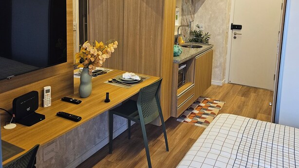 Een compacte, moderne kamer met een houten bureau, een flatscreen televisie, een klein kitchenette en een bed met een gestreept dekbed.