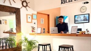 A reception area with a receptionist, a wooden desk, two black stools, a potted plant, and a wall clock.