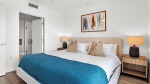 Ein Schlafzimmer mit einem großen Bett, zwei Nachttischen, einer zur Toilette führenden Tür und einem an der Wand hängenden abstrakten Gemälde.
