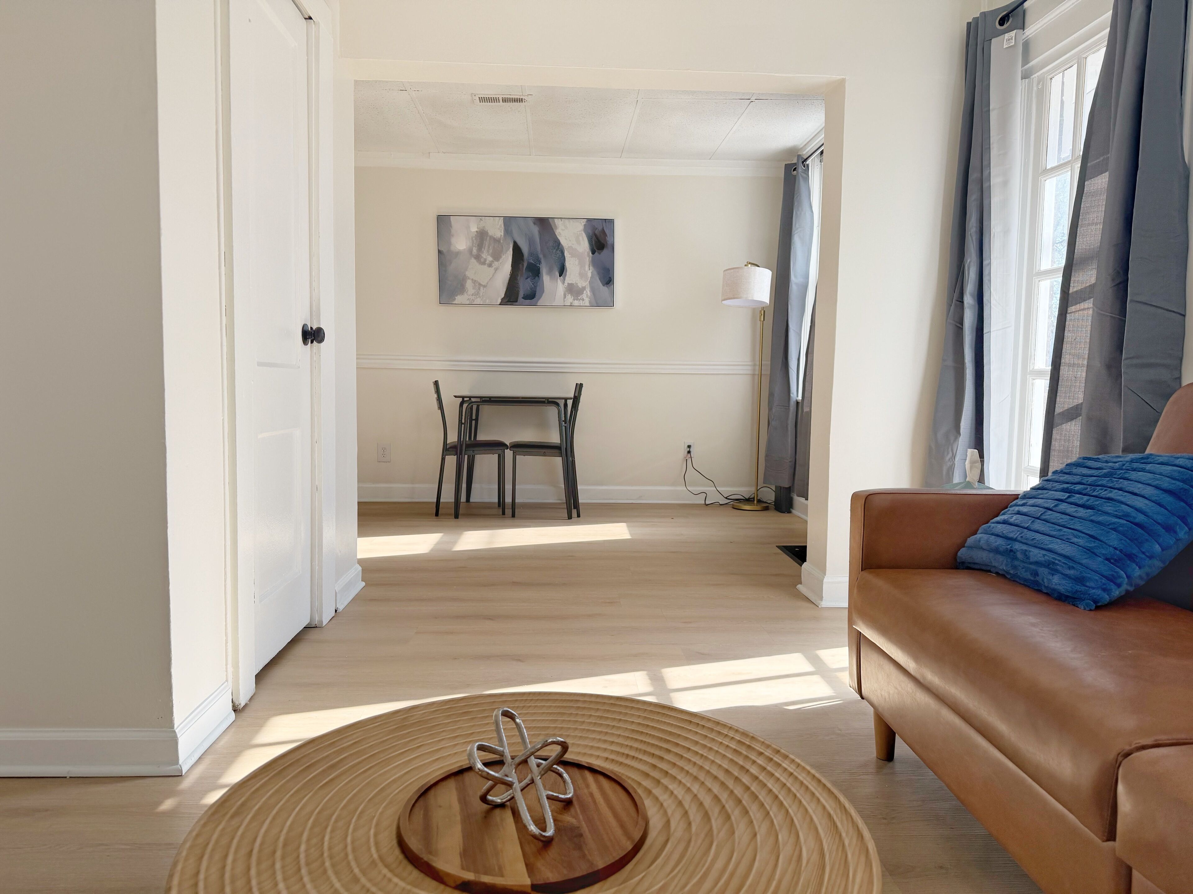 Een woonkamer met een bruine leren bank, een ronde houten salontafel met een metalen sculptuur, en een eethoek met een tafel en stoelen.