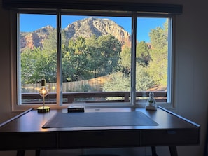 Ein Schreibtisch mit Lampe, Buch und einer kleinen Statue, mit Blick auf eine Berglandschaft durch ein großes Fenster.