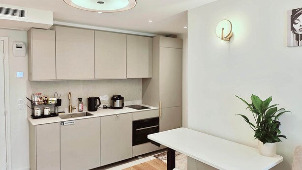 A modern kitchen with white cabinets, a built-in oven, a coffee maker, and a small dining table with a potted plant. - Paris- Issy les moulineaux - Appartement the King (Issy-les-Moulineaux)